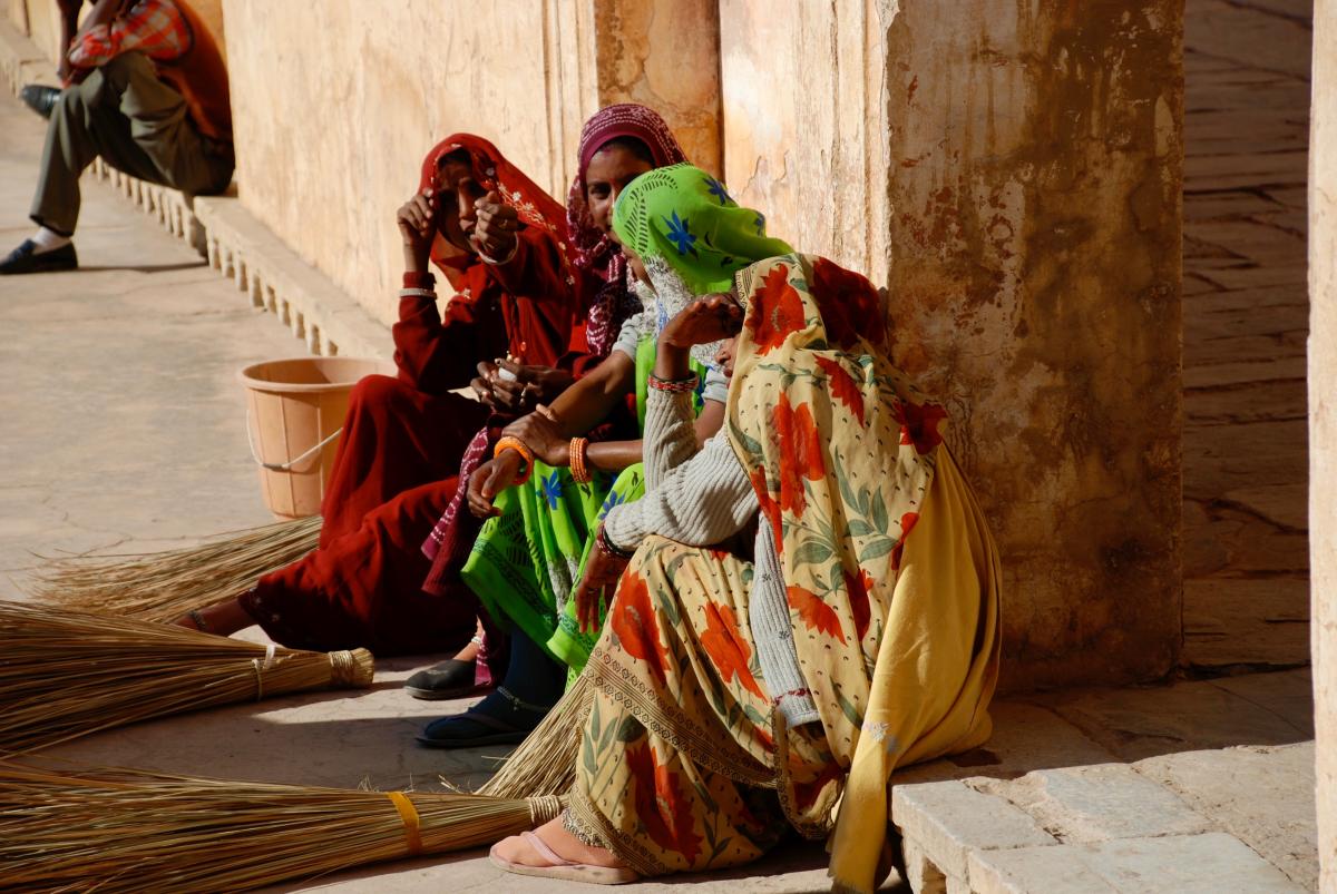 Three women in headscarves sit on the side of the street. They are talking to eachother. Two are facing the camera and one is not.