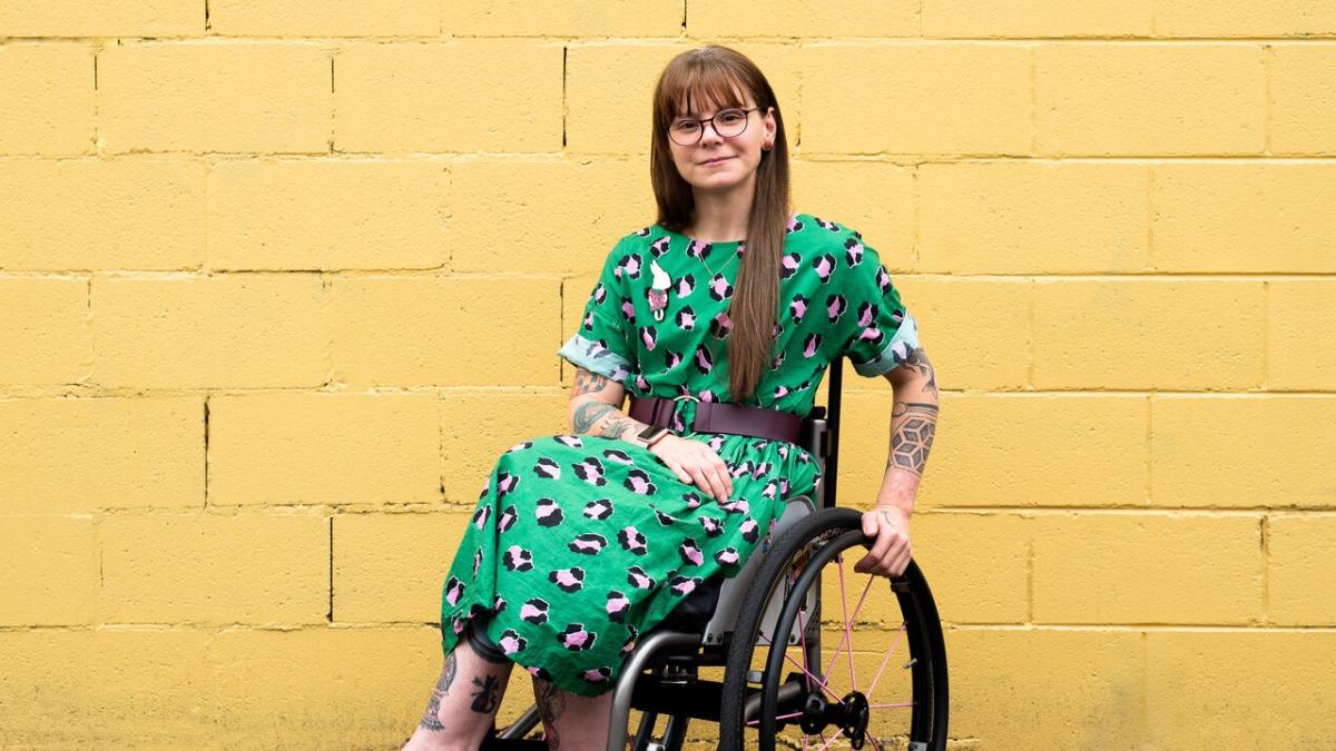 A woman in a wheelchair sits in front of a yellow brick wall. She has long brown hair, glasses, amazing tattoos and is wearing a green dress. 