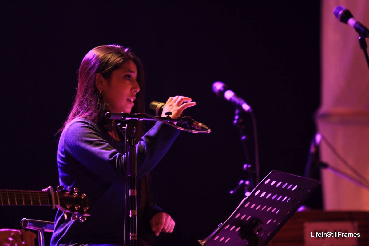 A young woman on stage singing into a micrphone. She is lit with purpke and blue lights.