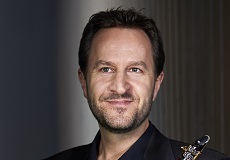 A man with short brown hair and a short brown beard smiles with his lips closed. He is holding a clarinet.