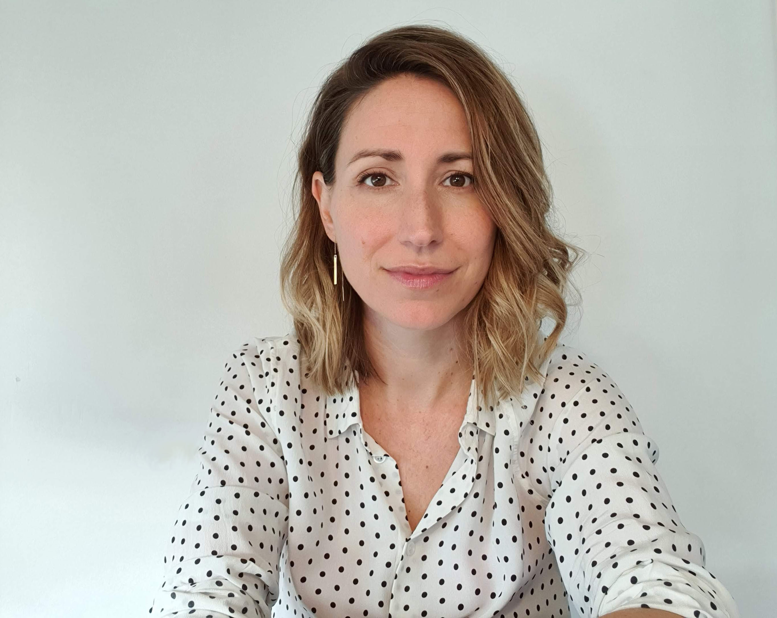 Isabelle is looking at the camera, giving a soft smile. She is a woman with shoulder length, curly blonde hair. She is wearing a white button up shirt with black polka dots. 