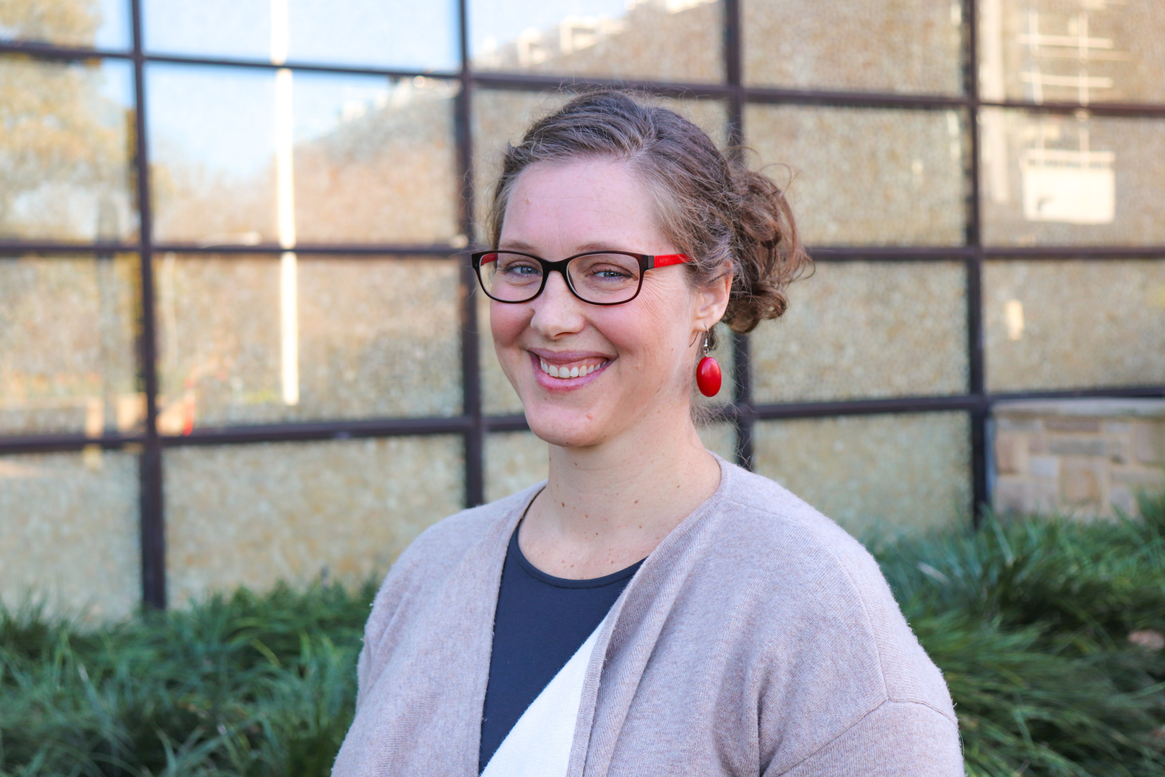 Lisa is smiling at the camera. She has brown hair tied up in a bun. She is wearing glasses and red earrings. 