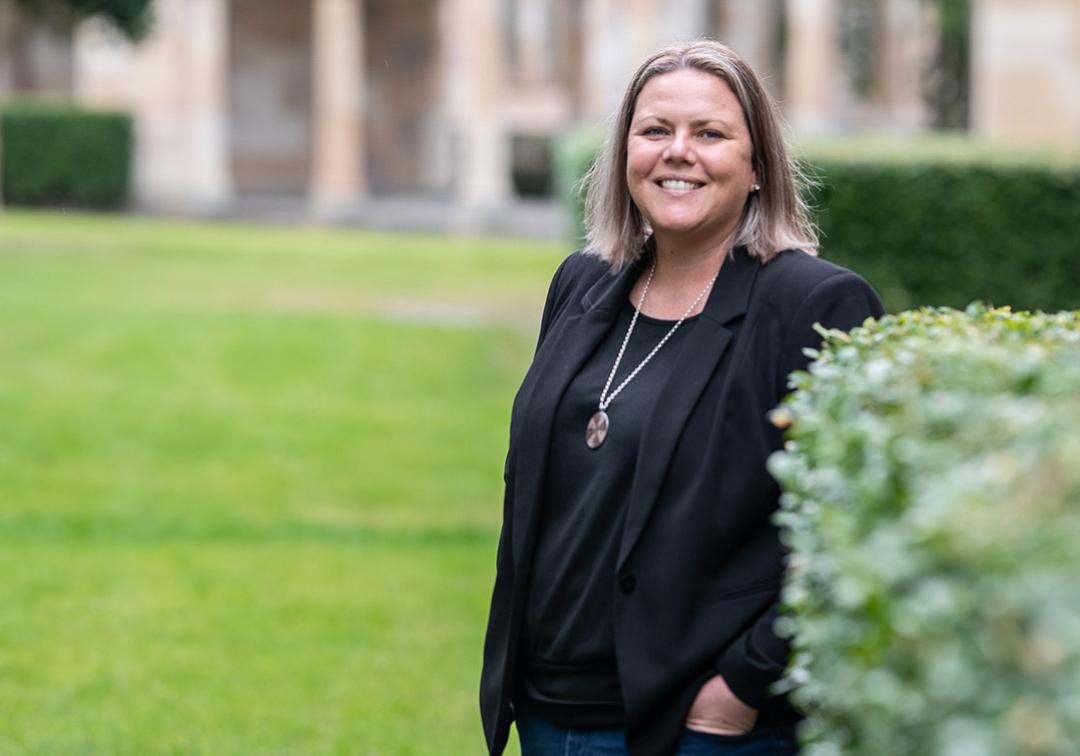 Marnee is outside in a green courtyard, standing next to a hedge. She is smiling at the camera. She has shoulder length, straight brown hair with blonde highlights. She is wearing a black blazer and a long necklace. 