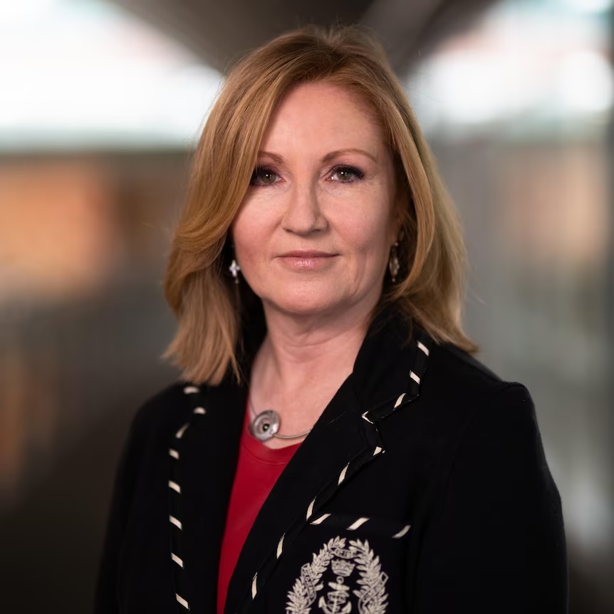 Headshot of Adele Ferguson, with strawberry blonde, straight hair, a black blazer with a red shirt underneath
