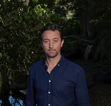 Dr Andrew Dansie stands outdoors in front of dense greenery and trees, wearing a dark blue button-up shirt. 