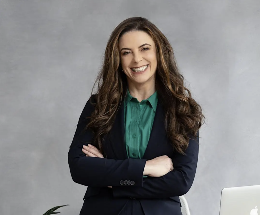 A smiling woman with long, wavy brown hair stands with her arms crossed, wearing a dark blazer over a green blouse against a gray backdrop.