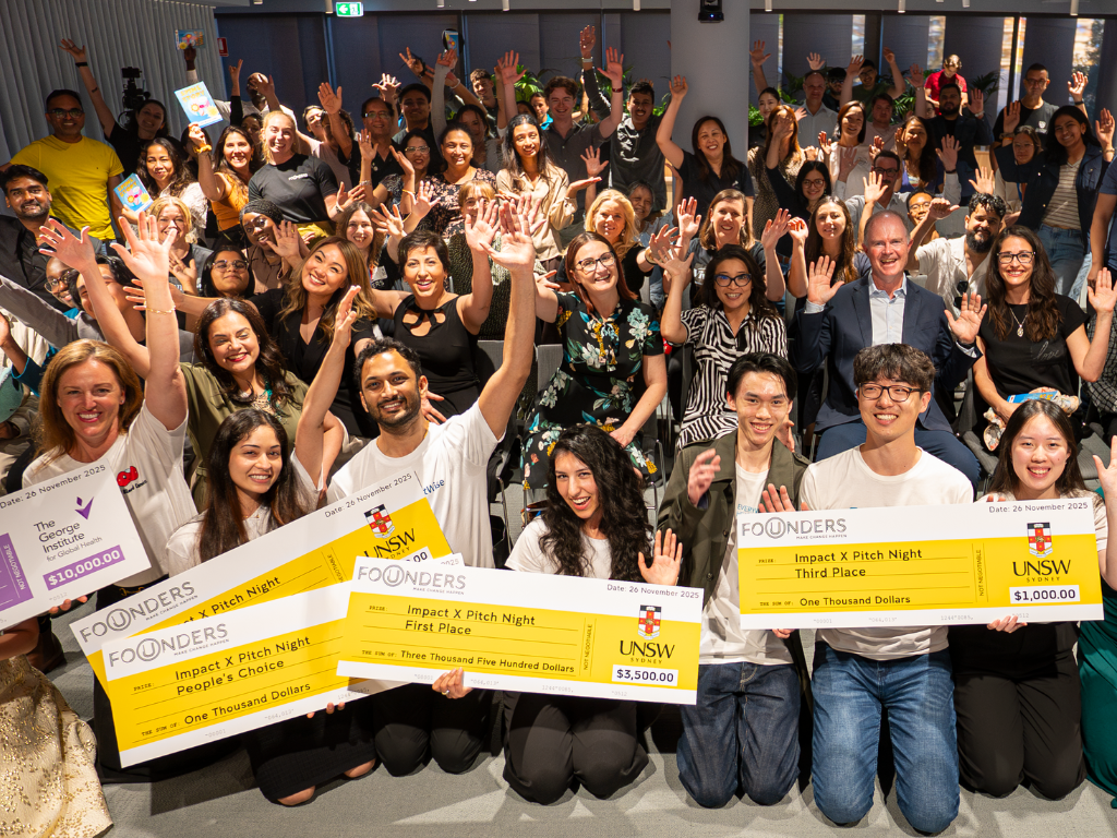 Group of audience members raising their hand in celebration. In front of the group, are eight founders holding four cheque prizes.