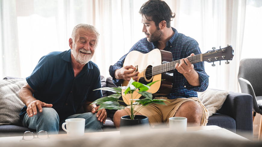 An older man sitting on a couch next to a young man who is playing a guitar