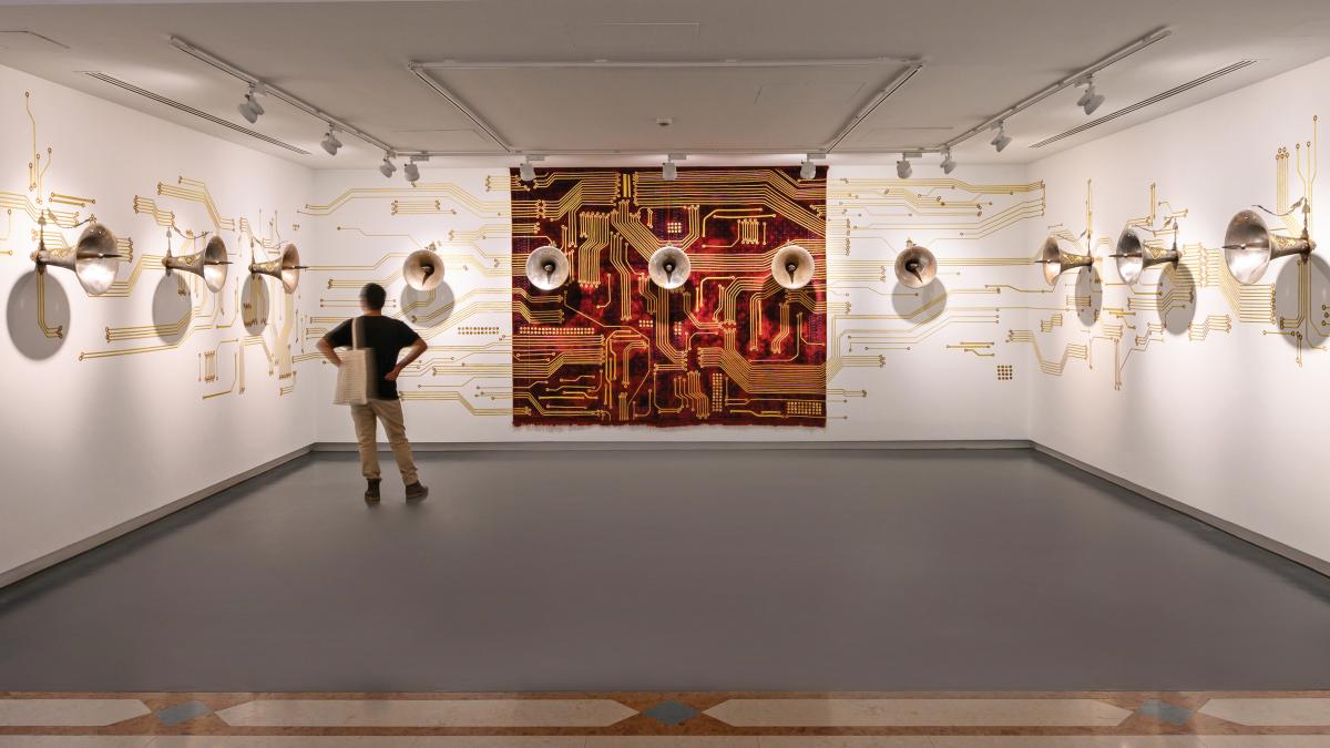 A person stands in front of an artwork installation by Khadim Ali comprising tapestry, gold thread, and ornamental steel speakerphones.
