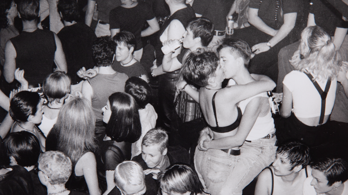 Archival image of two women in a party crowd kissing. 