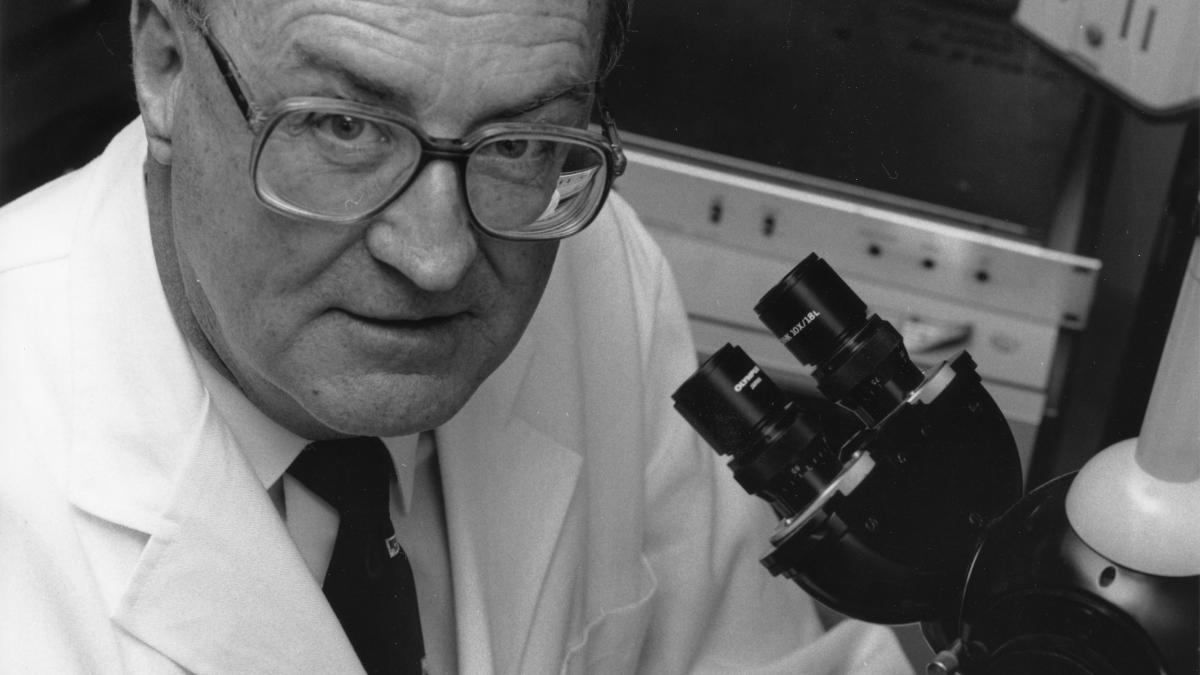 Black and white photo of a professor in a lab coat looking through a microscope