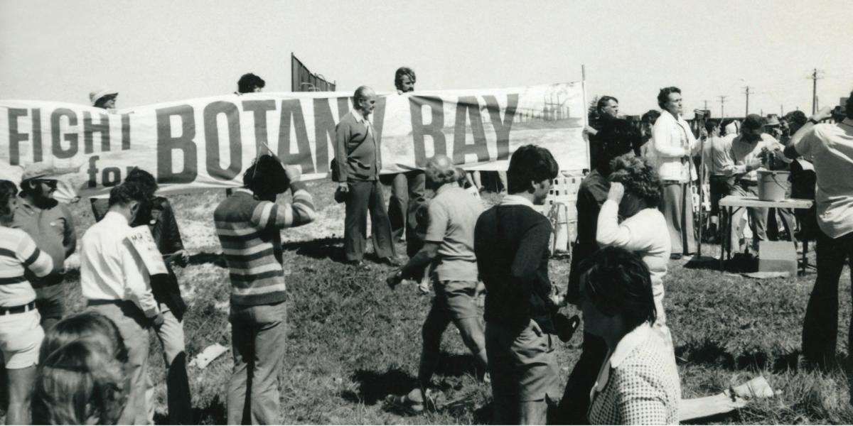 Black and white image of people gathered in protest with a banner in the background that reads ' fight for Botany Bay'