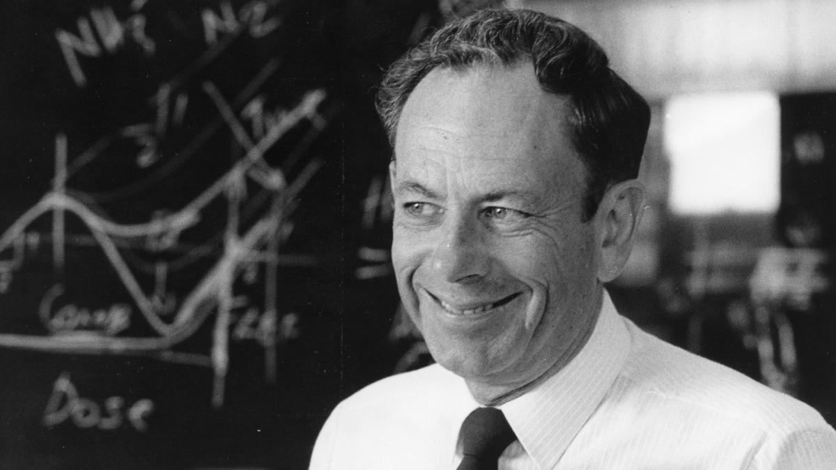 Black and white photo of a man in a suit with a kind smile. Behind him is a blackboard with mathematical equations. 