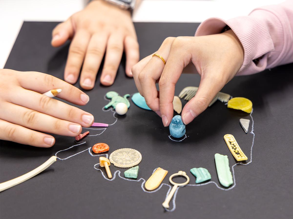 "Two hands arranging small, colorful objects on a black surface, including a key, coin, and trinkets. One hand holds a blue object, the other an white pencil."