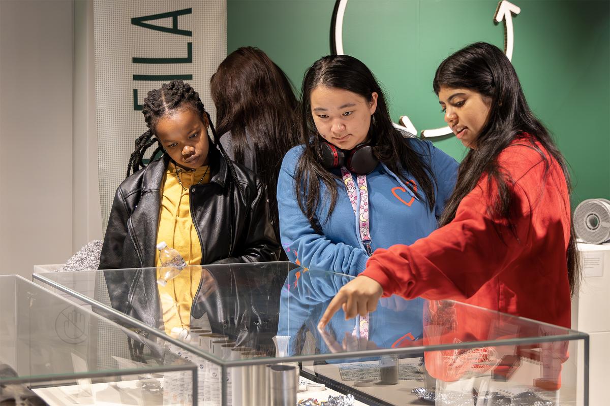 Three people gathered around a glass vitrine, one woman pointing at the display within an exhibition space.