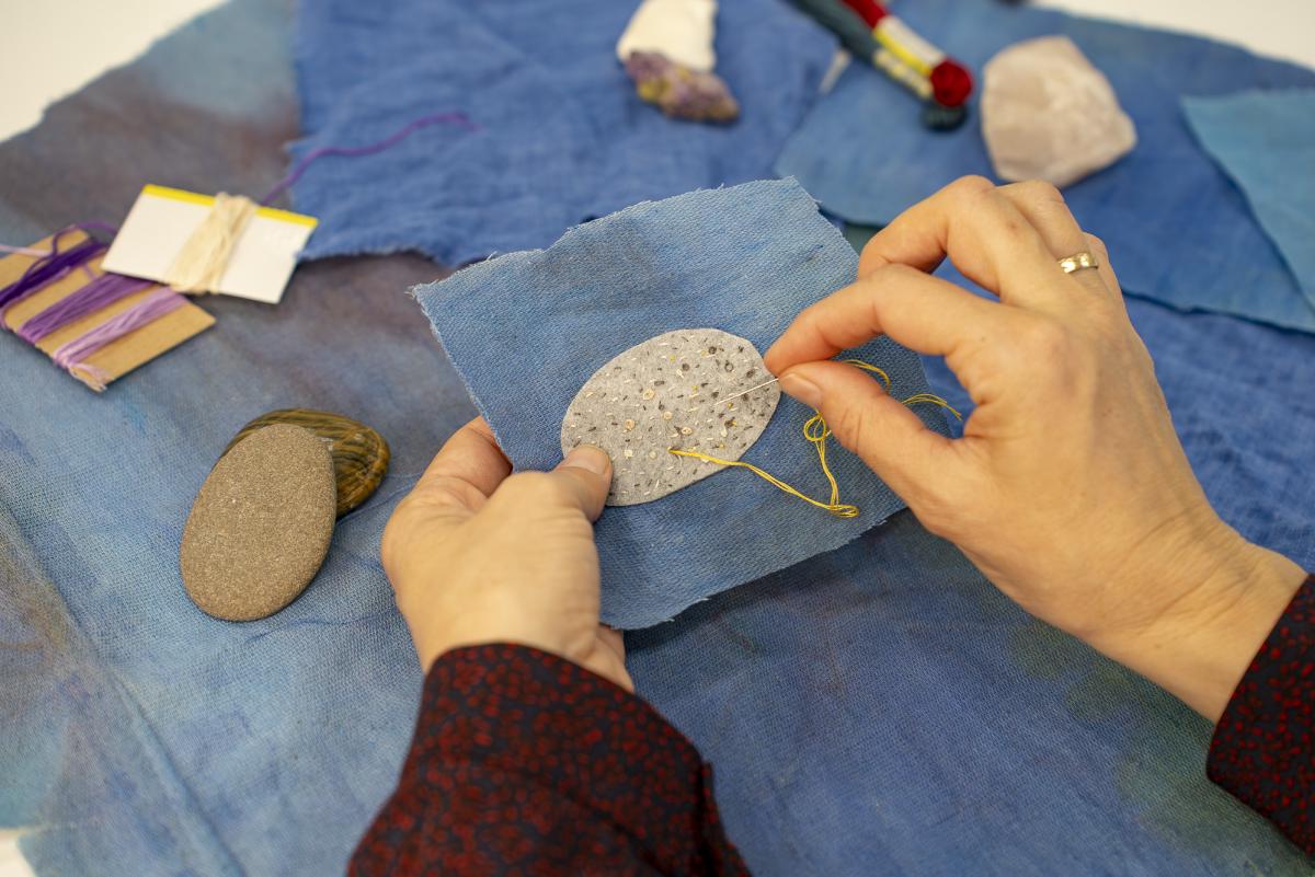 Hands stitching a grey oval fabric onto a blue square with rocks and sewing materials in the background.