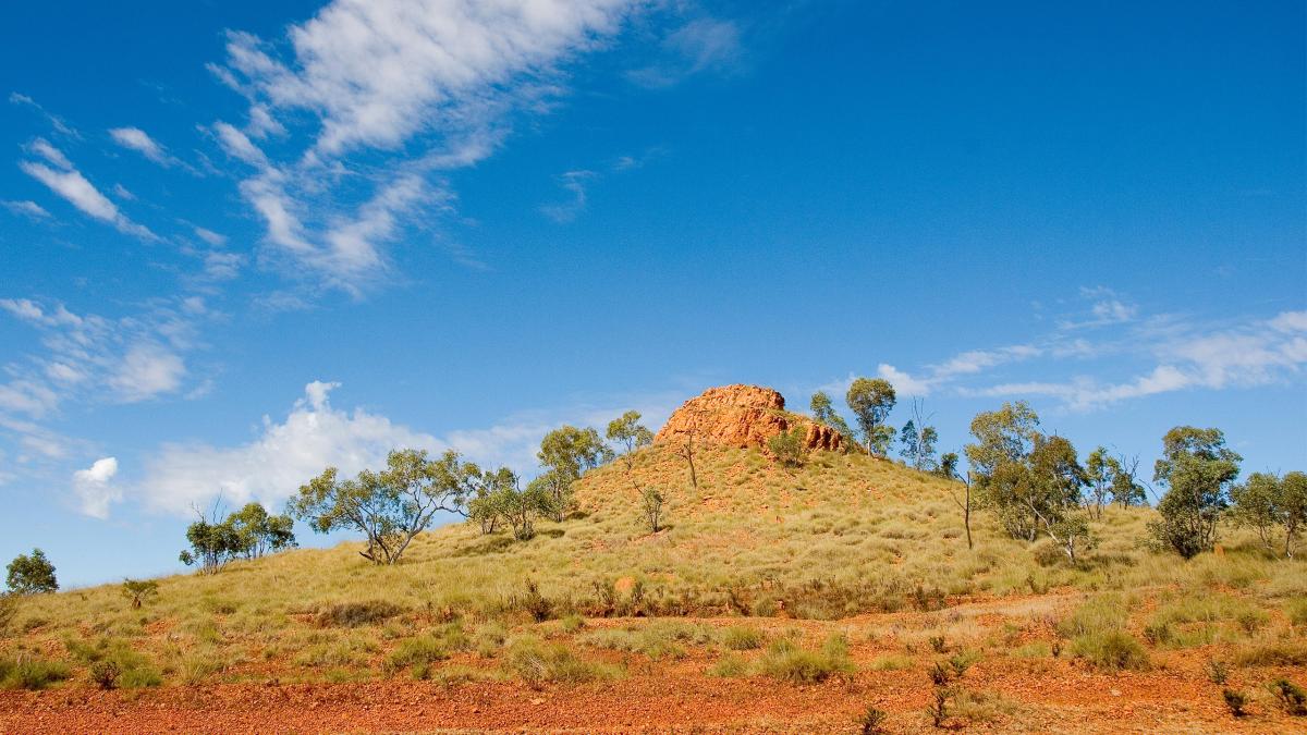 A semi-arid Australian landscape with reddish-brown earth and a cluster of large rocks on top of a hill surrounded by green scrub. Behind the rocks, the sky is a deep blue with scattered clouds. 