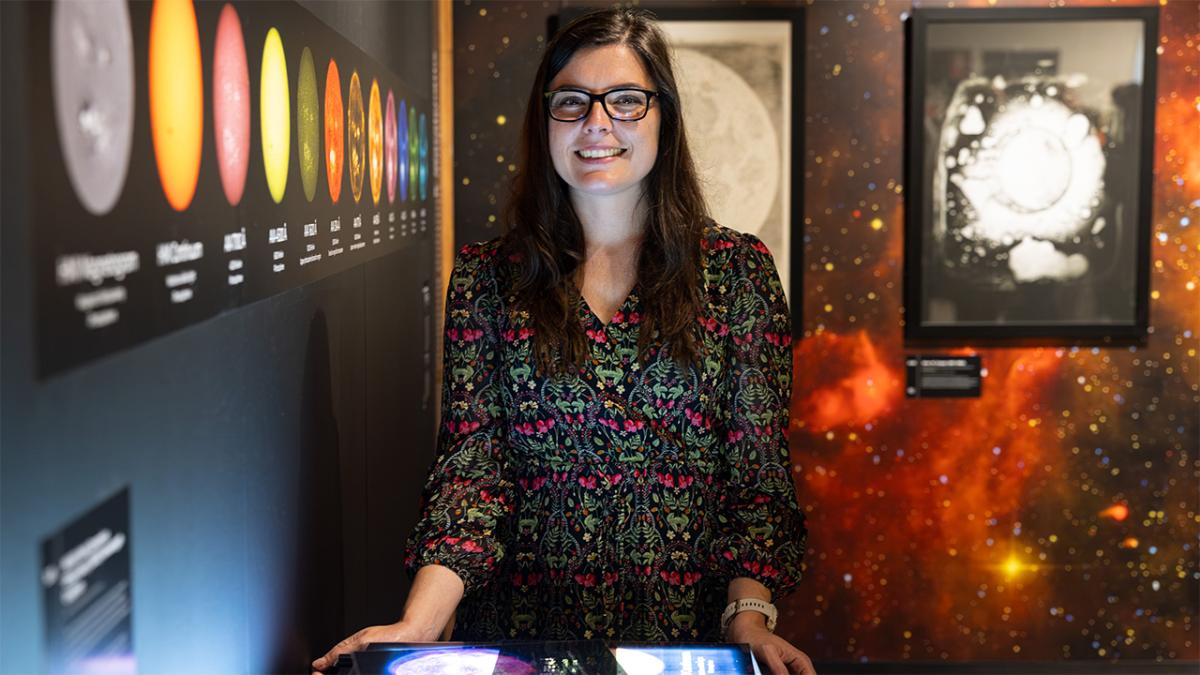 An astrophysicist stands smiling behind a glowing astronomical display, with a sequence of colourful solar icons on the wall beside her.